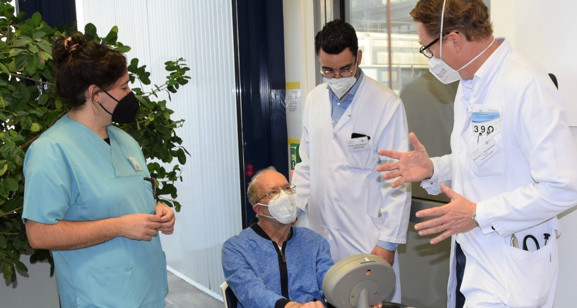 Mann sitzt auf einem Stuhl und tretet in Pedale, ähnlich zu einem Standfahrrad. Zwei Mitarbeiter und eine Mitarbeiterin des Klinikums stehen um ihn herum.