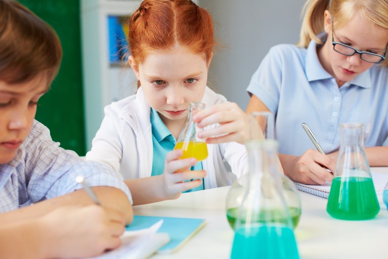 Eine junge Schülerin betrachtet einen Kolben mit gelber Flüssigkeit, weitere Schüler machen sich Notizen